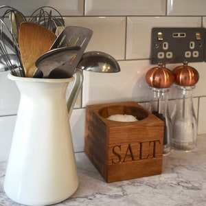 May include: A white ceramic jug with a handle, filled with kitchen utensils, including a wooden spoon, a whisk, and a metal spatula.  A wooden salt container with the word "SALT" engraved on the front sits on a grey countertop.  Two glass salt and pepper shakers with copper tops are behind the salt container.