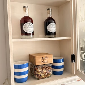May include: A white cabinet with shelves displays two bottles of liquor, two blue and white striped mugs, and a jar of nuts with a wooden lid that reads "Dad's Nuts". Wine glasses are visible above.