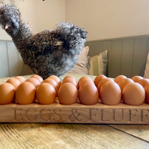May include: A wooden egg holder with the text "EGGS & FLUFF" engraved on the front. The holder is filled with brown eggs. A fluffy gray chicken is standing behind the holder.