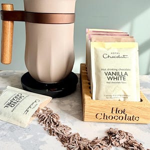 May include: A hot chocolate set featuring a beige mug with a wooden handle and a copper-colored band. Packets of hot chocolate, including vanilla white and salted caramel, are displayed with chocolate flakes. A wooden holder with "Hot Chocolate" is also present.