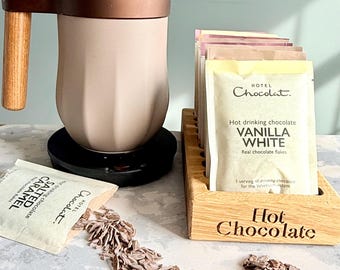 May include: A hot chocolate set featuring a beige mug with a wooden handle and a copper-colored band. Packets of hot chocolate, including vanilla white and salted caramel, are displayed with chocolate flakes. A wooden holder with "Hot Chocolate" is also present.