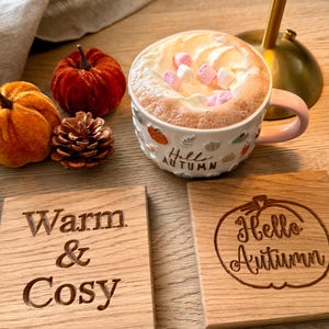 May include: A ceramic mug filled with a beverage topped with whipped cream and pink marshmallows, featuring the text "Hello AUTUMN". Two square wooden coasters are present, one engraved with "Warm & Cosy" and the other with "Hello Autumn".