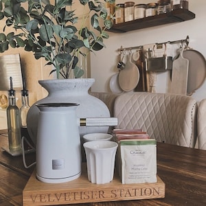May include: A white electric milk frother with a gold handle sits on a wooden cutting board with the words "Velvet  Sister Station" engraved on it.  There are four white ceramic mugs and a box of Hazelnut Mocha Latte coffee pods on the board.