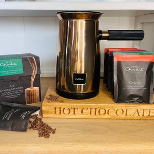 May include: A gold-colored milk frother sits on a wooden tray with the words "HOT CHOCOLATE" engraved on the front.  The tray holds several packets of hot chocolate mix, including flavors like Mint Drinking Chocolate, Orange, and Caramel.  The packets are from the brand "Hotel Chocolat."
