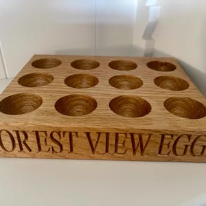 May include: A wooden egg holder with 12 individual compartments. The holder is made of light brown wood and has the words "FOREST VIEW EGGS" engraved on the front.