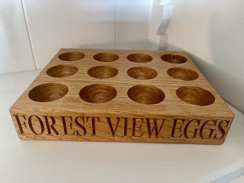 May include: A wooden egg holder with 12 individual compartments. The holder is made of light brown wood and has the words "FOREST VIEW EGGS" engraved on the front.