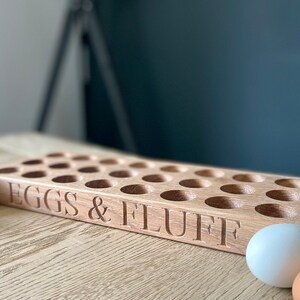 May include: A wooden egg holder with 24 slots, labeled "EGGS & FLUFF".  The holder is made of light brown wood and is sitting on a wooden table. There are several eggs on the table, including brown and white eggs.
