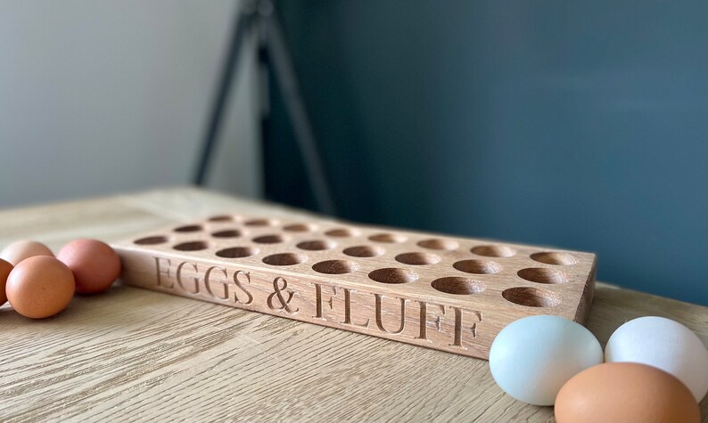 May include: A wooden egg holder with 24 slots, labeled "EGGS & FLUFF".  The holder is made of light brown wood and is sitting on a wooden table. There are several eggs on the table, including brown and white eggs.