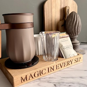 Könnte beinhalten: Ein Holzserviertablett mit dem eingravierten Text „MAGIC IN EVERY SIP“ enthält eine braune und beige Kaffeemaschine, ein klares Glas, eine weiße Tasse und Teebeutel. Ein Holzschneidebrett und dekorative Gegenstände befinden sich im Hintergrund.