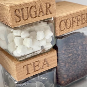 May include: Three glass jars with wooden lids labelled "Sugar", "Coffee", and "Tea". The jars are filled with sugar cubes, coffee beans, and tea bags.