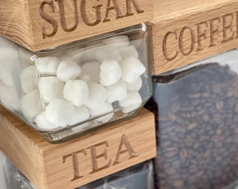 May include: Three glass jars with wooden lids labeled "Sugar", "Coffee", and "Tea". The jars are filled with sugar cubes, coffee beans, and tea bags.
