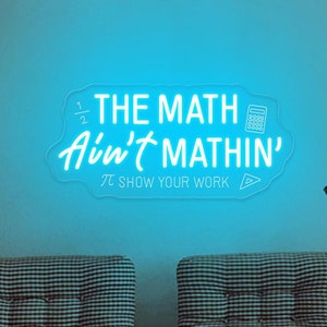 Op de afbeelding: Een blauw neonbord met de tekst "The Math Ain't Mathin' 1/2 π Show Your Work" met een rekenmachine-icoon en een driehoek-icoon.