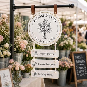 Può includere: Un'insegna rotonda bianca appesa con cinghie in pelle marrone, recante il testo Bloom & Stem Floral Studio e l'illustrazione di un bouquet. Sotto ci sono tre cartelli rettangolari bianchi con le scritte Fresh Flowers, Custom Bouquets e Wedding Florals.