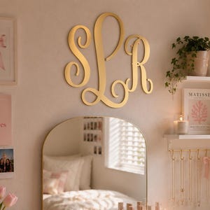 Puede incluir: Un escritorio blanco con un espejo dorado arqueado, un portátil, organizadores de maquillaje de acrílico transparente, un jarrón con tulipanes rosas, un soporte para joyas dorado y un letrero de pared con el monograma dorado SLR.