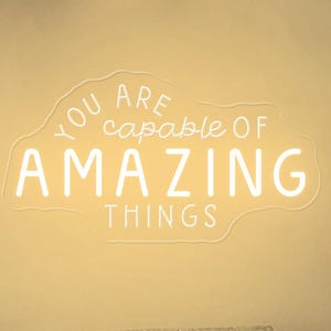 Puede incluir: Un salón con un sofá, cojines y un letrero de neón que dice "You are capable of AMAZING THINGS". La habitación está decorada con grabados artísticos, una lámpara de pared con forma de rosquilla y un jarrón con flores.