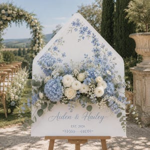 Op de afbeelding: Een wit, envelopvormig trouwbord met blauwe en witte bloemstukken, waaronder hortensia's en rozen. Het bord toont de tekst 'Aiden & Hailey' in blauw handschrift, gevolgd door 'EST. 2026' en een kleine blauwe afbeelding van bladeren.