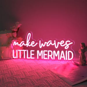 May include: Pink neon sign with the text "make waves little mermaid" on a white background.