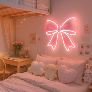 Op de afbeelding: Een roze neon boogvormige lichtarmatuur hangt boven een bed met roze beddengoed en decoratieve kussens. Een bureau met een stoel en bloemen in vazen zijn zichtbaar. De kamer heeft een zachte, pastelkleurige esthetiek.