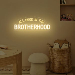 Könnte beinhalten: Ein weißes Neon-Schild mit dem Text "ALL GOOD IN THE BROTHERHOOD" in Wolkenform. Das Schild ist an einer beigefarbenen Wand montiert.