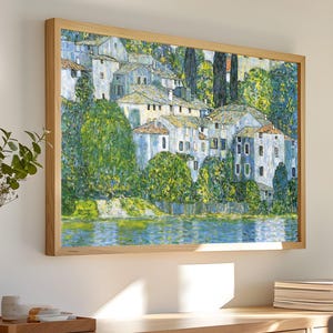May include: A framed painting of a village by a lake. The painting is done in a style reminiscent of impressionism, with visible brushstrokes and a focus on light and color. The village is depicted in shades of blue and white, while the lake is a vibrant blue. The surrounding trees are a lush green.