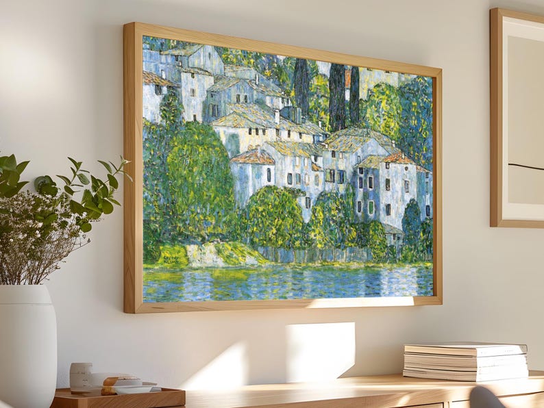 May include: A framed painting of a village by a lake. The painting is done in a style reminiscent of impressionism, with visible brushstrokes and a focus on light and color. The village is depicted in shades of blue and white, while the lake is a vibrant blue. The surrounding trees are a lush green.