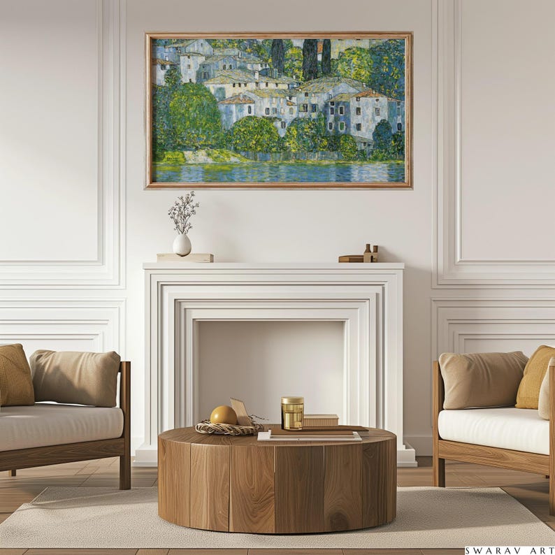 May include: A framed painting of a village with white buildings and green trees, hanging on a white wall above a fireplace in a living room. The fireplace has a white mantel and a white surround. There is a wooden coffee table in front of the fireplace with a wicker basket, a gold jar, and a book on top.