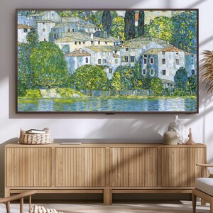 May include: A framed painting of a village with white buildings and green trees, set against a blue sky and water. The painting is hanging on a wall above a wooden cabinet.