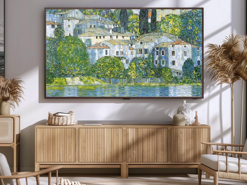 May include: A framed painting of a village with white buildings and green trees, set against a blue sky and water. The painting is hanging on a wall above a wooden cabinet.