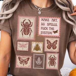 May include: Brown t-shirt featuring a collage of postage stamp designs with insect illustrations and the text "MAKE ART, DO SPELLS, FUCK THE SYSTEM!". The stamps are in shades of brown, pink, and beige.