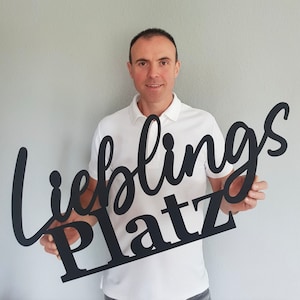 Könnte beinhalten: Ein schwarzes Holzschild mit den deutschen Worten "Lieblings Platz" in einer Schreibschrift. Das Schild wird von einer Person in einem weißen Poloshirt vor grauem Hintergrund gehalten. Das Schild ist ein dekorativer Artikel.