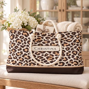 May include: A tote bag featuring a brown and black leopard print pattern with a solid dark brown base. The bag has thick white rope handles and a rectangular label on the front with the name CHRISTINA printed in black capital letters.