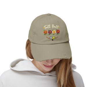 Peut inclure: Une casquette de baseball kaki à l'aspect usé, ornée de l'inscription Fall Ball en lettres blanches, accompagnée de quatre illustrations de tournesols colorés et de deux raquettes de pickleball croisées bleues et jaunes sur le devant.