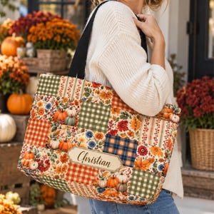 Op de afbeelding: Een rechthoekige draagtas met een patchworkontwerp van herfstpatronen, waaronder pompoenen, bloemen en ruitjes. De tas heeft zwarte schouderbanden en een centraal rechthoekig label met de naam Christian erop gedrukt.