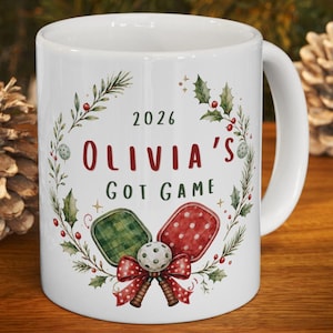 May include: A white ceramic mug featuring a festive wreath design with holly leaves and red berries. The text reads 2026 OLIVIA'S GOT GAME. Below the text are two pickleball paddles, one green plaid and one red polka dot, tied with a red bow.