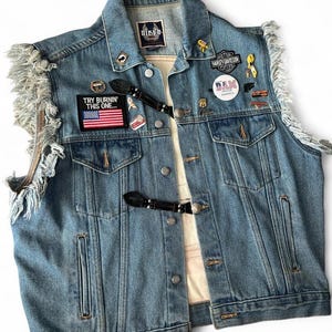 May include: A light blue denim vest with frayed edges and various pins and patches. The vest features a black leather buckle closure and the text "TRY BURNIN' THIS ONE..." on a patch with an American flag.
