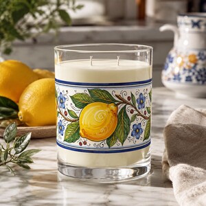 Peut inclure: Une bougie en verre transparent avec un intérieur en cire crémeuse. La bougie présente une bande décorative avec un motif de citron et de fleurs en jaune, bleu et vert. La bougie est à côté de citrons frais et d'un pichet décoratif.