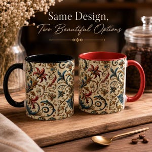 May include: Two ceramic mugs with a floral design. One mug has a black handle and interior, the other has a red handle and interior. Both mugs feature the same cream-colored background with red, blue, and gold floral patterns. The mugs are on a wooden surface.