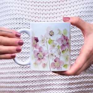 May include: White ceramic mug with a floral design featuring pink and white flowers. The mug is being held by a person's hands.