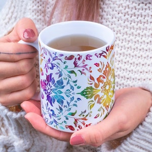 May include: A white ceramic mug with a colorful floral pattern. The mug is filled with a light brown liquid.