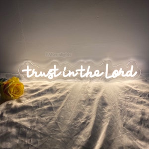 Può includere: Un cartello al neon bianco con le parole "trust in the Lord" in un carattere corsivo. Il cartello è su una superficie di tessuto bianco con una rosa gialla in primo piano.