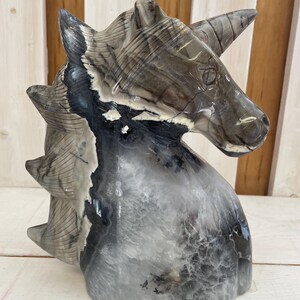 Puede incluir: Una escultura de unicornio tallada en madera petrificada. La escultura es de color gris claro con vetas negras y blancas. El unicornio tiene un cuerno largo y puntiagudo y una melena fluida.