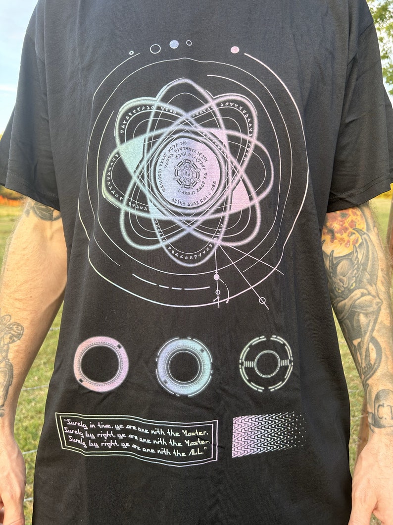 May include: Black t-shirt with a graphic design of a stylized atom with a circle in the center. The atom is surrounded by a series of concentric circles with text in a circular pattern. The text reads "Surely in time, ye are one with the Master. Surely by right, ye are one with the Master. Surely by right, ye are one with the ALL."