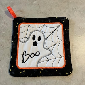 May include: Square Halloween pot holder featuring a white ghost with black eyes and the word "Boo". The design is set against a white background with a spiderweb pattern. It has a black border with yellow accents and an orange trim. A red ribbon is attached.