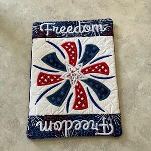 May include: Quilted rectangular item with a patriotic design. Features a central starburst pattern in red, white, and blue, with the word "Freedom" embroidered in white script at the top and bottom. The background is white with a diamond quilt pattern.