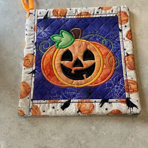 May include: A square quilted potholder featuring a central orange jack-o'-lantern appliqué on a dark blue background with a spiderweb pattern. The border is a light fabric printed with orange pumpkins, black crows, and stars, with an orange hanging loop.