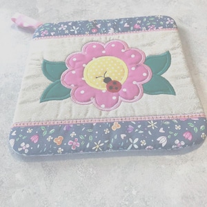 May include: A quilted pot holder featuring a large pink flower with white polka dots and a yellow center, a small ladybug, and green leaves. The edges are trimmed with a floral pattern in blue and pink, and a pink loop for hanging.