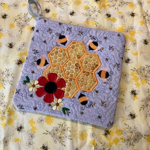 May include: A quilted lavender pot holder featuring a honeycomb design with embroidered bees. A large red flower with a black center and small white flowers are also embroidered on the pot holder. A light blue ribbon is attached for hanging.