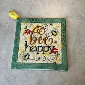 May include: Quilted square pot holder with the words "bee happy" embroidered in brown thread. The center has a floral pattern with bees and flowers. The border is green, and a yellow loop is attached for hanging.