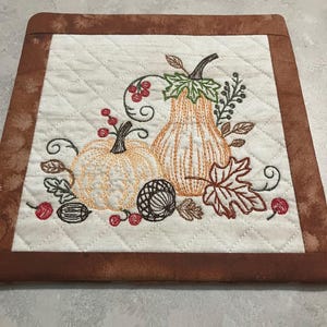 May include: A quilted, square, off-white fabric with a brown border features an embroidered autumn design. Two orange pumpkins, an acorn, and fall foliage are stitched in shades of brown, green, and red. The fabric has a diamond pattern.
