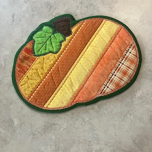 May include: A quilted fabric pumpkin-shaped mat featuring vertical strips of fabric in various shades of yellow, orange, and brown with different patterns. It includes a green fabric leaf and a brown stem, all bordered by a dark green edge.